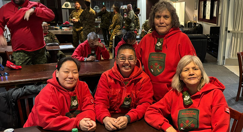 Gathered around the table are four smiling Rangers, each wearing the bright red Canadian Ranger hoodie. In the background are other Rangers and people in army camouflage uniforms.