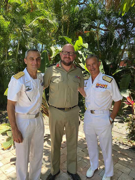 A man in a khaki military uniform stands between two men in Naval whites. The man in the middle is Eric Andresen. He is taller than the other two men, is wearing black rimmed glasses and has a short brown beard. The other two men are both clean shaven with short grey hair. The three stand arm in arm, with palm trees behind them.