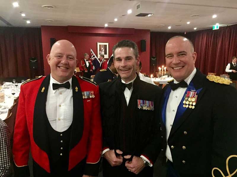 Three men in formal military dress uniforms smile for the camera during a mess dinner. Andresen is wearing a red and black formal uniform jacket, with military medals displayed, also a white dress shirt and a black bow tie. The other two men are wearing black dress uniforms with medals and a white shirt and black bow tie.