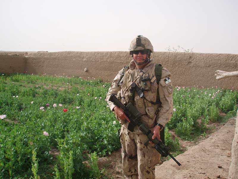 A man in desert camouflage and helmet carrying a large rifle. He is standing in front of a flower garden.