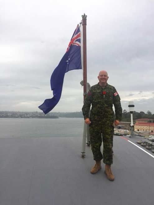 A man in military fatigues stands smiling in front of a large Australian flag. He is standing on the deck of a naval ship with the Sydney Harbour in the background.
