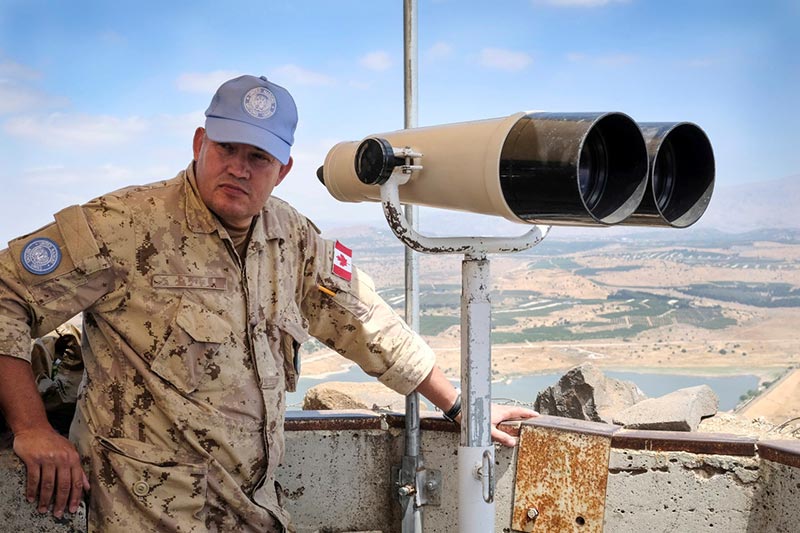 A man wearing khakis and a UN peacekeepers ballcap stands behind a large set of binoculars. He has a Canada flag patch on his left shoulder and is overlooking a desert area.