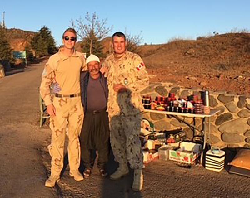 Two men in khakis stand with their arms around a smaller, older man with a moustache and a white cap. Behind them is a stone wall and a table full of bottles of honey.