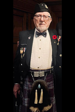 George Brewster, plus âgé, pose pour une photo où il est vêtu d’un kilt, d’un nœud papillon et d’un sporran assortis. Il porte des médailles militaires et un coquelicot rouge est épinglé sur son veston.