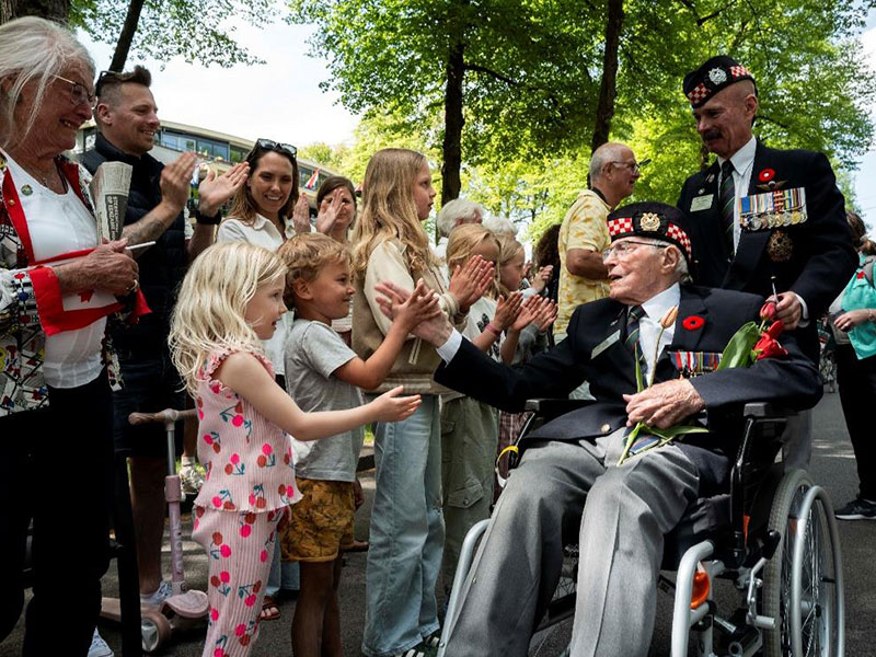 Un vétéran en uniforme tenant des fleurs rouges salue des enfants et d’autres participants lors d’un défilé. Des personnes rassemblées à proximité l’applaudissent lorsqu’ils se rencontrent.