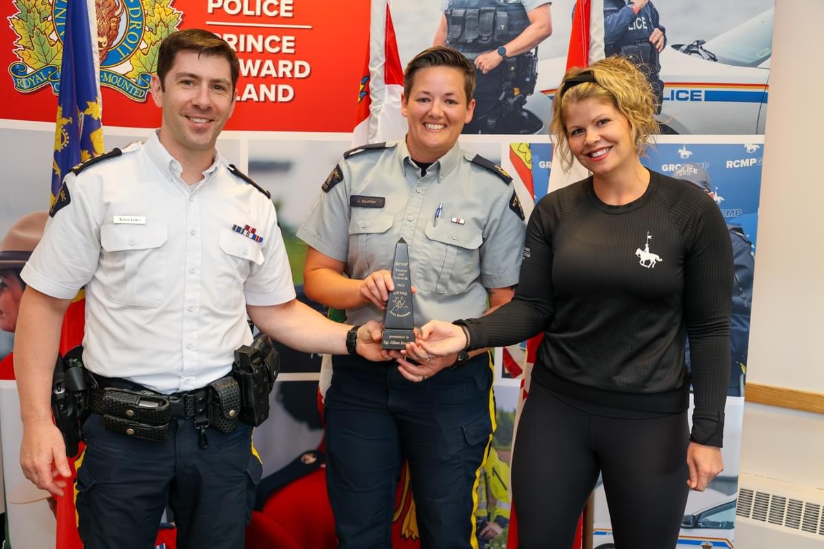 Boutilier is standing with two other people, she is in the middle. Chief Superintendent of the RCMP, Kevin Lewis is on her right. He is wearing a white uniform shirt with blue jeans. Elysia Atkinson is on her left. She is wearing a black shirt and pants, the shirt has a white horse on the left side of the shirt representing the musical ride. They are presenting her with an award. Jillian is wearing her general duty uniform and is smiling at the camera.