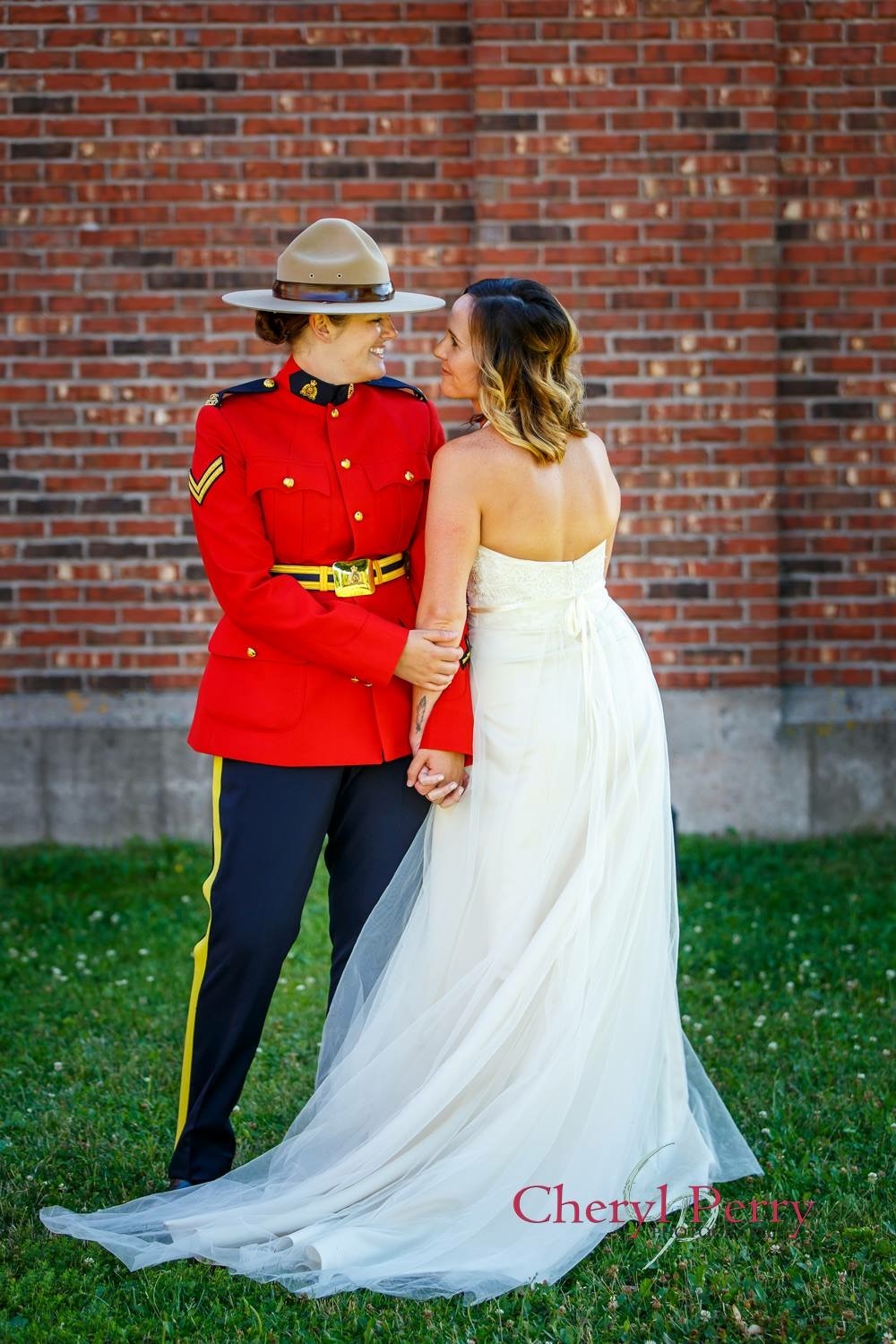 Boutilier and her wife are standing in front of a brick wall. They are side by side, but facing each other. They are holding hands and looking into each other’s eyes. Boutilier is in the RCMP Red Surge dress uniform and her wife, (Bethany Boutilier), is wearing a white, strapless wedding gown.