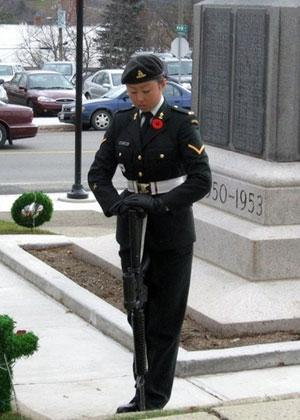 Une jeune femme chinoise en uniforme militaire se tient devant un cénotaphe, la tête inclinée et les mains posées sur un fusil. Elle porte un uniforme militaire et un coquelicot à son revers.