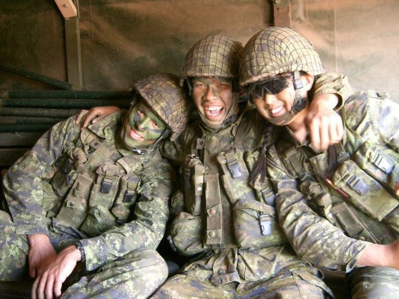 Trois jeunes soldates en tenue de combat de camouflage sont assises, souriantes, les bras autour des épaules les unes des autres. Deux portent du maquillage de camouflage et la troisième un casque de combat et des lunettes de soleil.