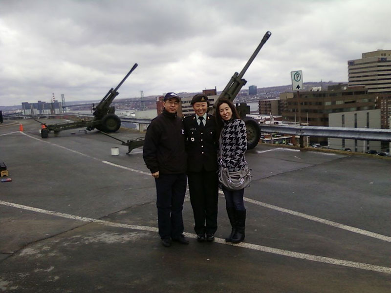 Une jeune femme chinoise en uniforme militaire se tient sur un quai devant deux canons militaires. À ses côtés, ses parents en tenue civile posent pour la photo.