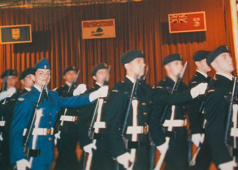 A group of soldiers in black military dress uniforms marching. One woman, in blue dress uniform, smiles at the camera.
