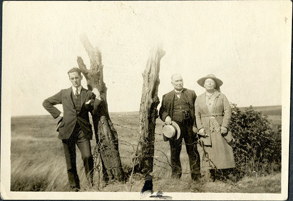 Photo en noir et blanc de trois adultes debout à l’extérieur près de troncs d’arbres brisés dans un champ.