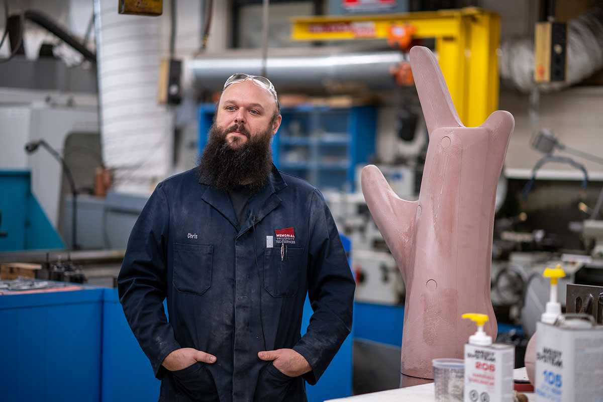 Une personne se tient dans un atelier à côté d’un grand modèle de tronc d’arbre rose, avec des machines et des outils en arrière-plan.