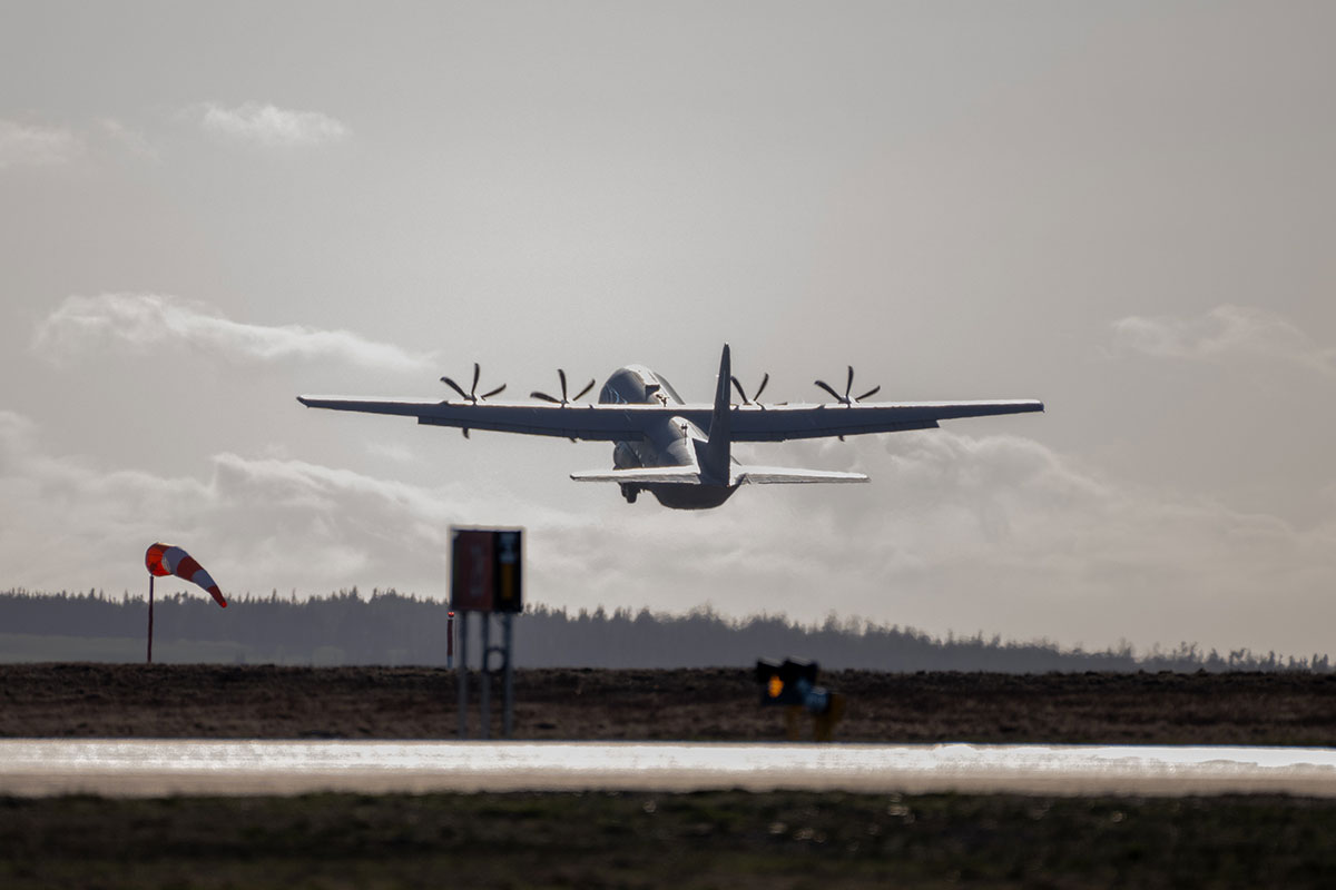 Un avion militaire décolle d’une piste, avec un indicateur de direction du vent et des nuages bas à l’arrière-plan.