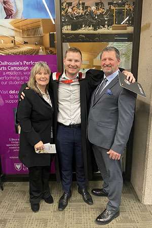 A man wearing a university graduation gown smiles with his arms around his parents on either side of him. They are standing in front of a Dalhousie University sign, and he is holding a graduation certificate.
