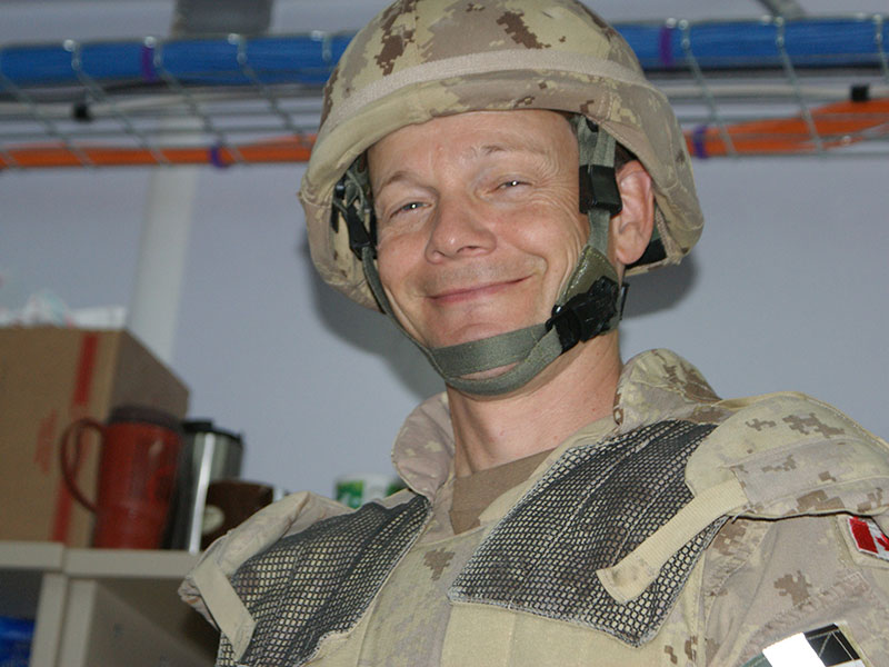 A soldier in desert fatigues and a helmet grins for a photo. There is a Canadian flag patch sewn on his shoulder.