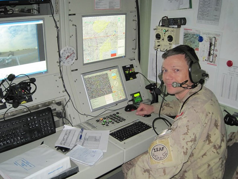 A soldier in desert fatigues sits at a desk with several computer monitors. He is wearing a headset with a microphone and turning over his left shoulder to smile at the camera.