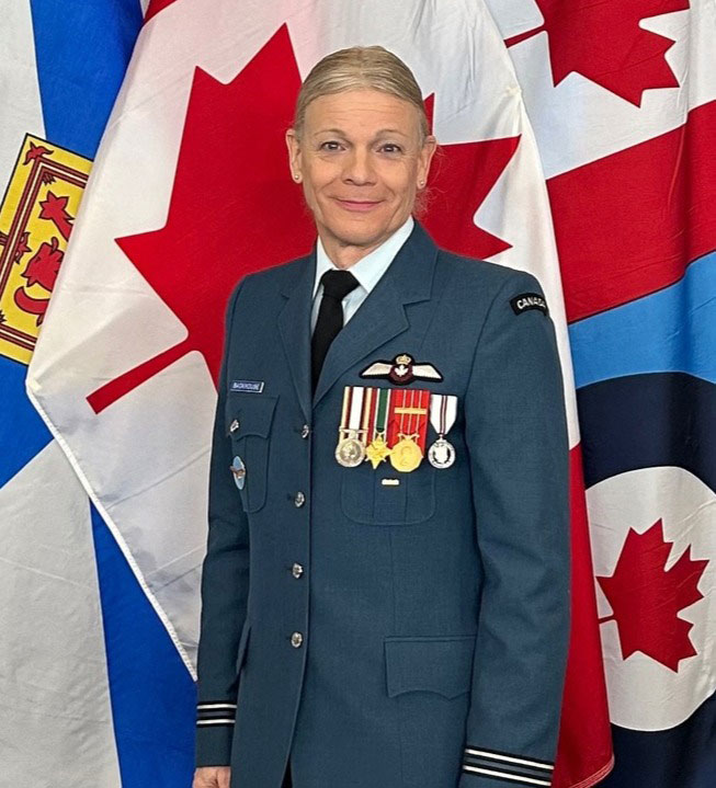 A woman in military dress uniform stands smiling in front of three flags; Nova Scotia, Canada and Royal Canadian Air Force. There are several military medals on her chest and her hands are at her sides.