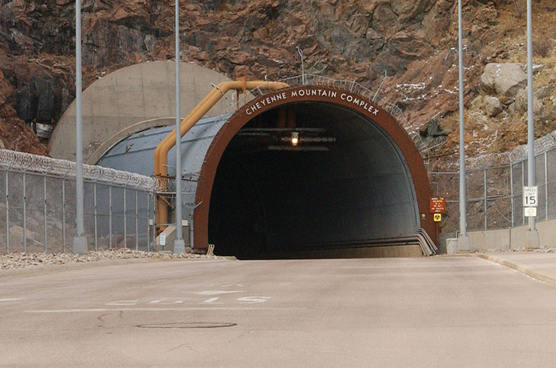 A grey road leads to a blue fortified vehicle tunnel into the base of a rocky mountain. Grey fencing topped with rolled barbed wire lines both sides of the road.