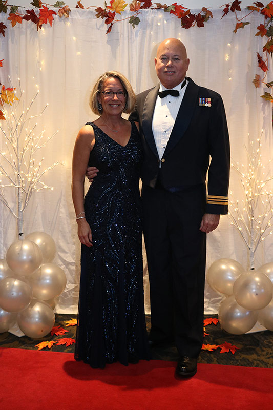 A Royal Canadian Air Force major in formal dress wearing his medals with his arm around his partner. She is wearing a long black dress. They are standing on a red carpet. The backdrop is a white curtain with a garland of orange, red and yellow maple leaves strung across the top. On each side of the couple is a tall white tree branch lit with white lights. A bunch of white shiny balloons is at the base of each branch.