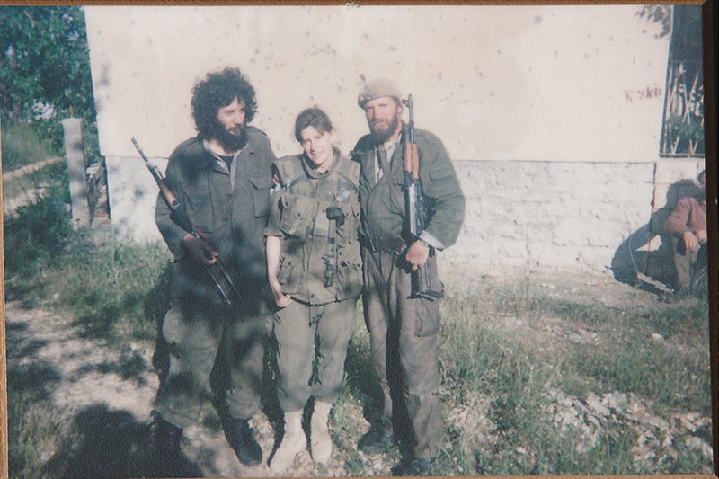 Sandra Perron stands between two men. All are wearing military fatigues. The two men look disheveled and are carrying firearms. They are standing in front of a beige and white wall outdoors. You can see trees and a fence in the back left corner.  