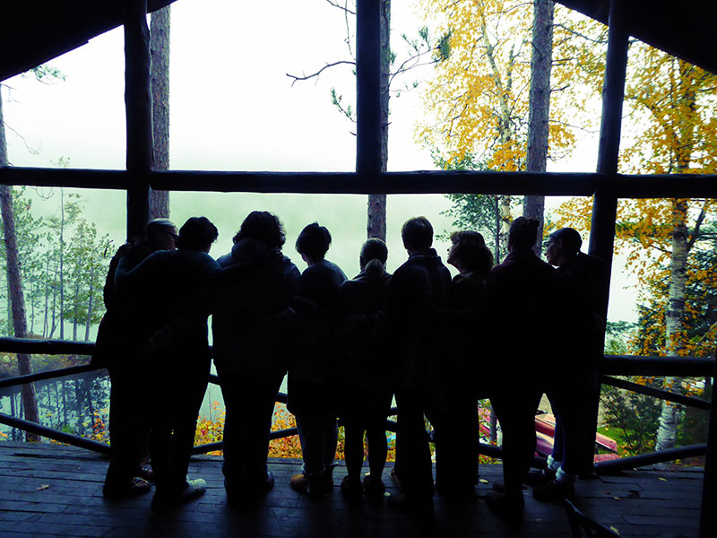 Nine women are standing in silhouette in front of a large window. They all have their arms around each other and are looking out toward a picturesque view of the forest.