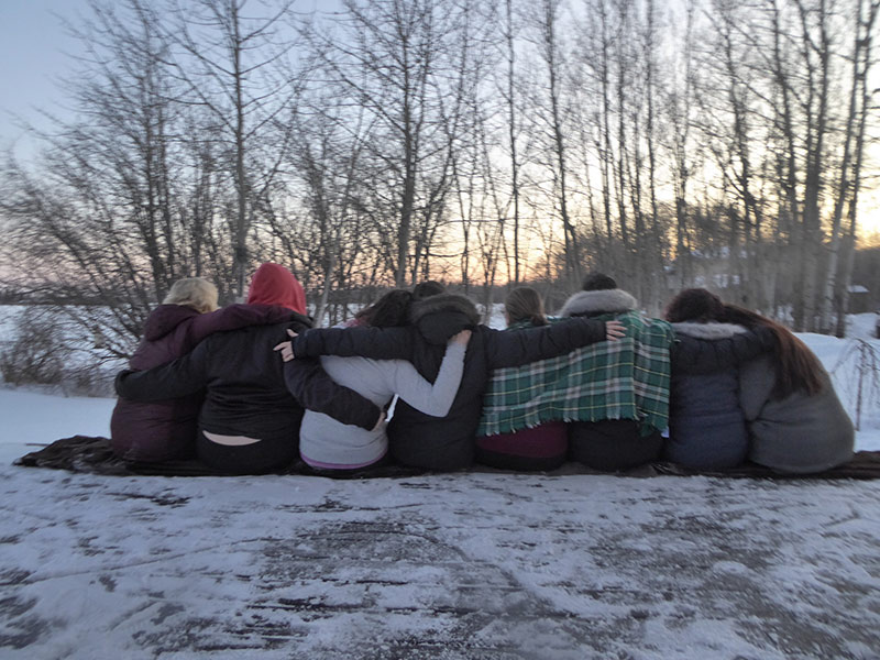 Eight women sit on the snowy ground. They are all arm in arm dressed in warm coats and winter blankets.