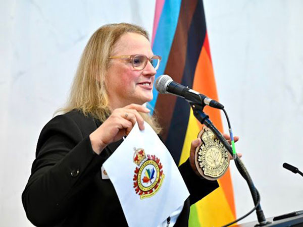Sherry Lundrigan speaking in front of a microphone, with a Pride flag behind her. She is holding a plaque and a paper with a rainbow-coloured maple leaf.