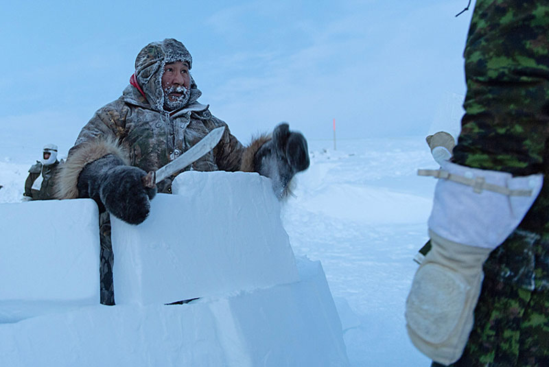 Solomon Awa talking to a Canadian Armed Forces member while standing behind blocks of snow. Solomon is holding a carving knife and wearing thick winter clothing.