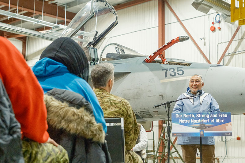 Solomon Awa standing at a podium giving a lecture to a crowd of people. A military jet is behind him.