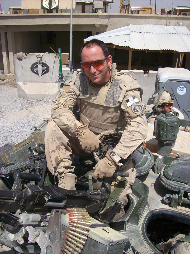 Steve Jourdain, vêtu d’un treillis militaire, est assis, entouré de munitions. Il sourit et porte des lunettes de soleil.