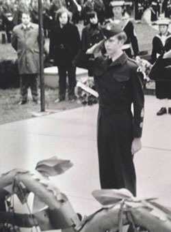 a black and white photo of a very young Steven Deschamps saluting. Steven is facing the camera and there are two Remembrance Day wreaths visible in the foreground. A crowd of civilians and Naval officers stand behind him.