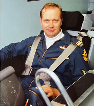a 20-something Steven Deschamps sits in the cockpit of an aircraft simulator. He is wearing an Air Force flying suit and is grinning at the camera.