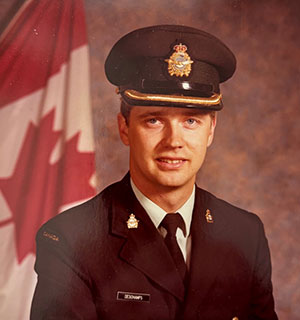 Steven Deschamps sits for a formal portrait. He is wearing a dark green military uniform with a black peaked cap with gold piping and a gold cap badge. He is posting with the Canadian flag behind his right shoulder.