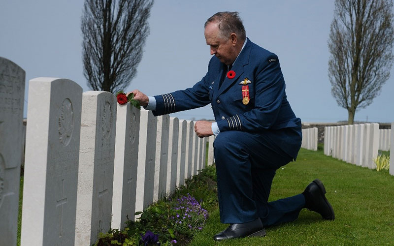 Steven Deschamps is down on one knee in front of a white headstone in Belgium. There are multiple rows of headstones lining the green space. He is wearing a blue Air Force uniform with a poppy on the lapel and black shoes. He is placing one red rose on top of the white headstone.
