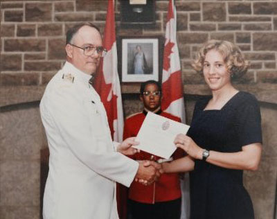 A young blonde woman in a black dress smiles for a photo accepting a certificated from an older man in a white military dress uniform.