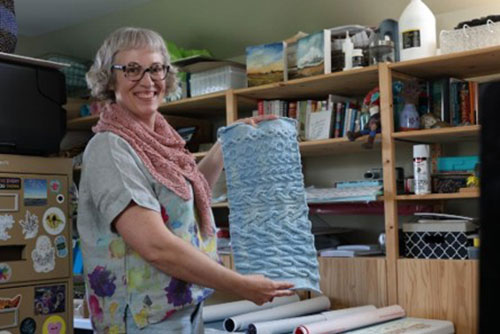A woman smiles holding a blue section of knitted yarn. She is wearing glasses, a pink scarf and a floral linen blouse.