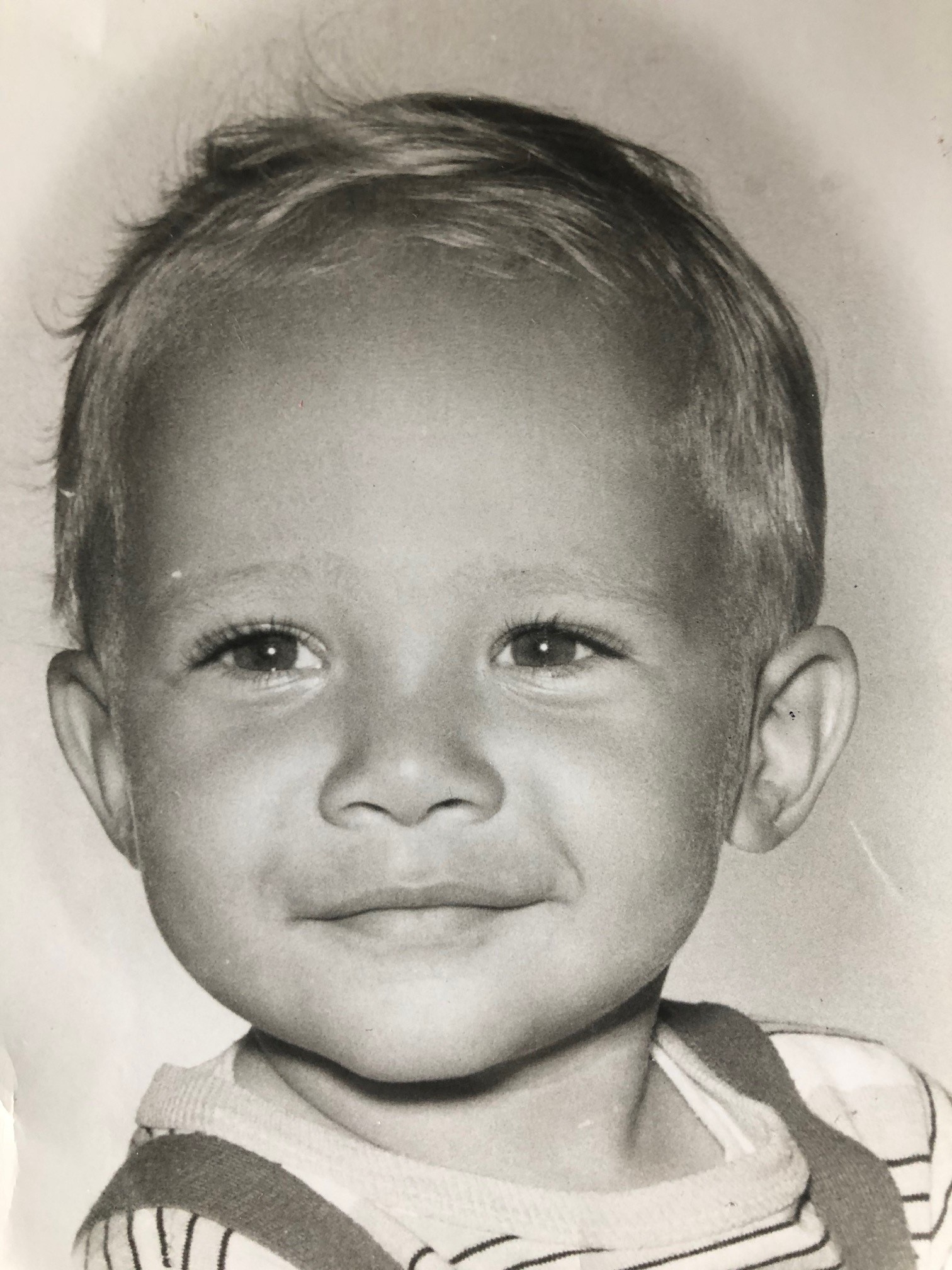 Une photo en noir et blanc de Treena Scurlock alors qu’elle était enfant. Elle porte un t-shirt rayé et des salopettes et sourit timidement à la caméra.