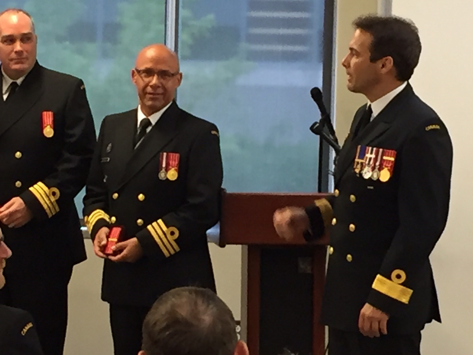 La capitaine de frégate Treena Scurlock, en uniforme noir de la Marine, reçoit la médaille CD1. Elle se tient entre deux autres officiers de la Marine et sourit à la caméra.