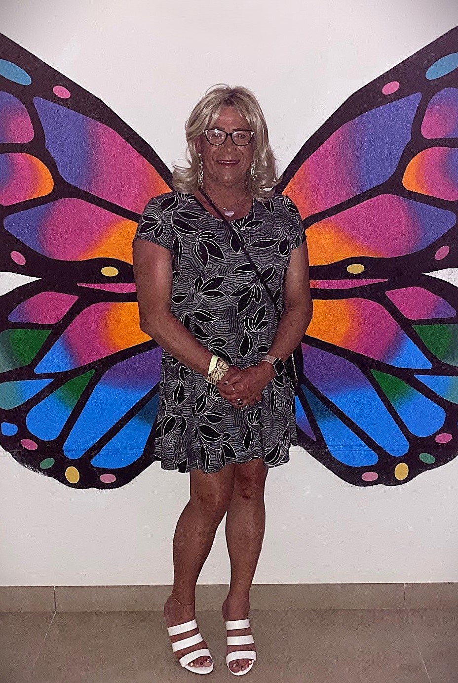 Treena Scurlock devant des ailes de papillon colorées. Elle porte une robe d’été noir et blanc, des sandales blanches, les mains jointes devant elle. Elle sourit à la caméra.