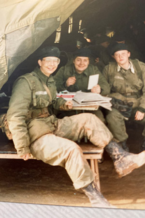 Three young women in army green fatigues and muddy combat boots sit at a picnic table inside a military tent, they are smiling at the camera.