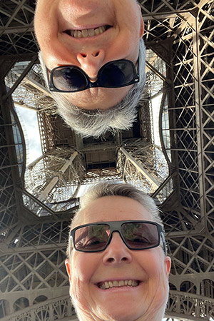 Two women with short grey hair and black sunglasses look down at the camera from underneath the Eiffel Tower.