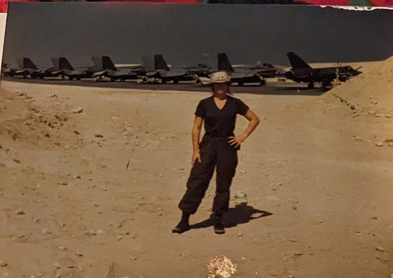 A woman stands in the desert sand, a row of military aircraft behind her. She is wearing fatigues, a beige wide brimmed sun hat and her left hand is on her hip.