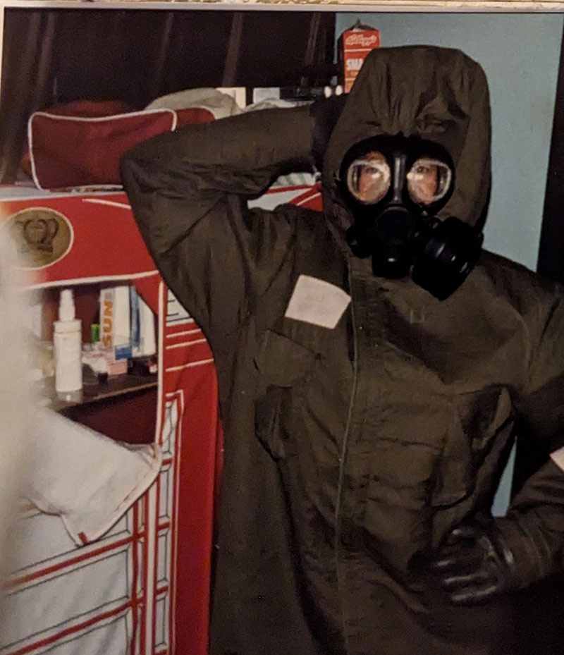 A woman wearing a gas mask and protective suit stands with one hand on her hip.