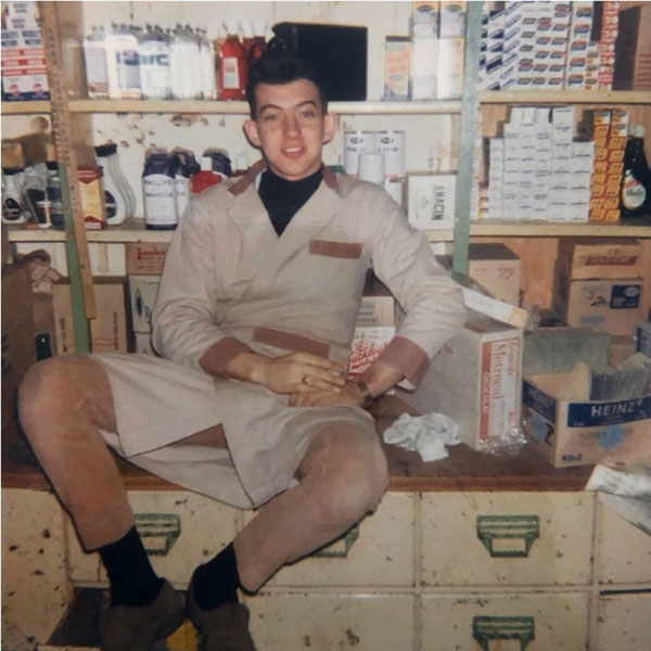 Wayne Davis, jeune homme. Il est assis à un comptoir, devant divers produits. Il porte une blouse beige et un pantalon marron.