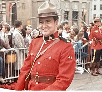 Plan moyen de Wayne Davis. Il sourit et ne regarde pas la caméra. Il porte la tunique rouge. Derrière lui, une foule est contenue de l’autre côté d’une clôture, et il y a un autre agent de la GRC devant la foule, qui se trouve en arrière de Wayne. 